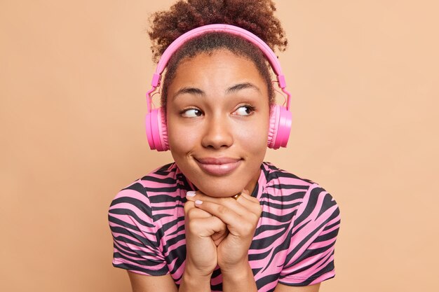 Portrait en gros plan d'une femme heureuse et réfléchie garde les mains sous le menton porte des écouteurs sur les oreilles écoute une piste audio ou une conférence de didacticiel concentrée de côté isolée sur un mur beige