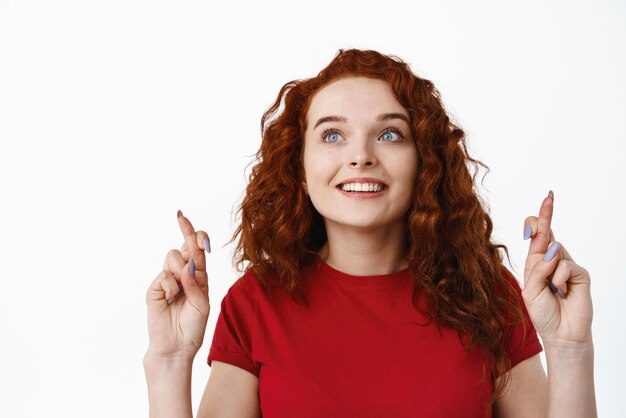 Portrait en gros plan d'une femme pleine d'espoir rousse aux cheveux bouclés croiser les doigts pour la bonne chance en regardant le logo du coin supérieur gauche avec un visage tenté veut quelque chose prier ou faire un vœu