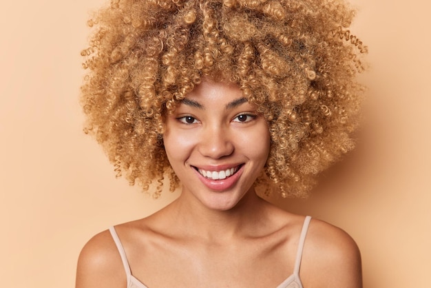 Portrait en gros plan d'une femme positive aux cheveux bouclés qui sourit doucement a une peau saine et soyeuse sans maquillage pose les épaules nues regarde directement la caméra isolée sur fond beige. Notion de beauté.