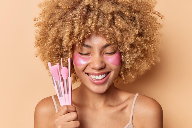 Portrait en gros plan d'une femme sincère positive aux cheveux bouclés qui sourit largement a des dents blanches applique des patchs de collagène sous les yeux pour réduire les rides tient des pinceaux cosmétiques pour appliquer le maquillage