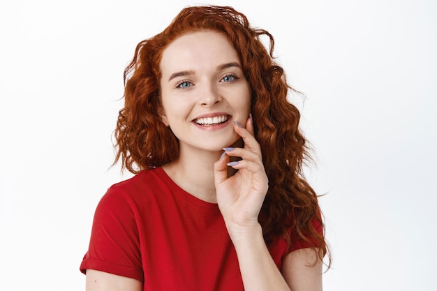 Photo gratuite portrait en gros plan d'une femme souriante et heureuse aux cheveux bouclés au gingembre, touchant une peau pâle, lisse et saine du bout des doigts, l'air gai et riant, mur blanc