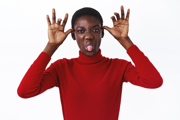 Portrait en gros plan d'une fille afro-américaine drôle et insouciante avec une coupe de cheveux courte et un col roulé rouge, montrant des grimaces