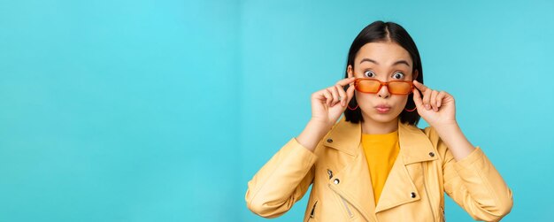 Portrait en gros plan d'une fille asiatique à lunettes de soleil à la recherche d'excitation heureuse et surprise sur le visage debout sur fond bleu
