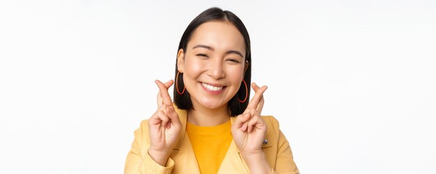 Portrait en gros plan d'une fille asiatique pleine d'espoir souhaitant croiser les doigts pour la bonne chance priant et souriant debout sur fond blanc Copier l'espace
