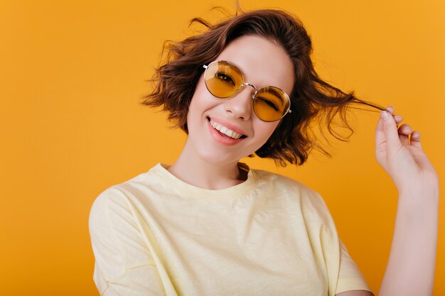 Portrait en gros plan d'une fille blanche insouciante joue avec des cheveux courts ondulés. Photo d'une femme européenne élégante avec un tatouage noir isolé sur un mur jaune.