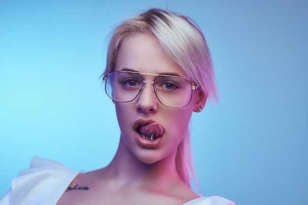 Portrait en gros plan d'une fille blonde passionnée portant des lunettes pose avec la langue qui sort en regardant la caméra. Isolé sur un fond bleu