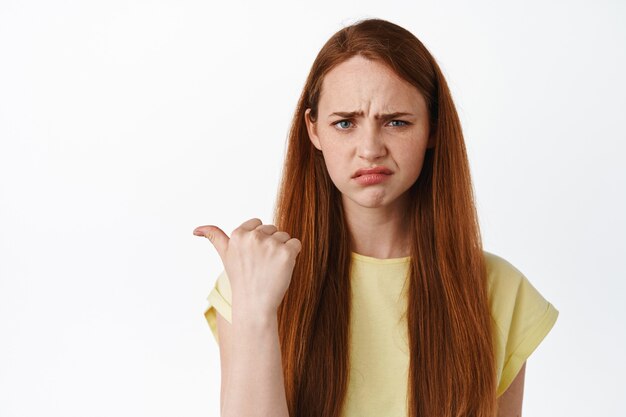 Portrait en gros plan d'une fille fronçant les sourcils triste et déçue, pointant vers la gauche quelque chose de déplaisant et de bouleversant, se plaignant, debout sur blanc