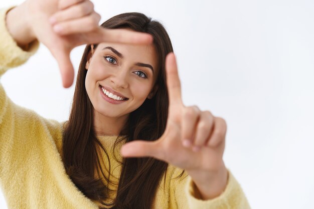 Portrait en gros plan fille inspirée heureuse rêveuse souriant joyeusement, regardez la caméra à travers les cadres des doigts comme si elle imaginait un moment, imaginez une scène ou essayez de capturer une superbe photo, un mur blanc debout