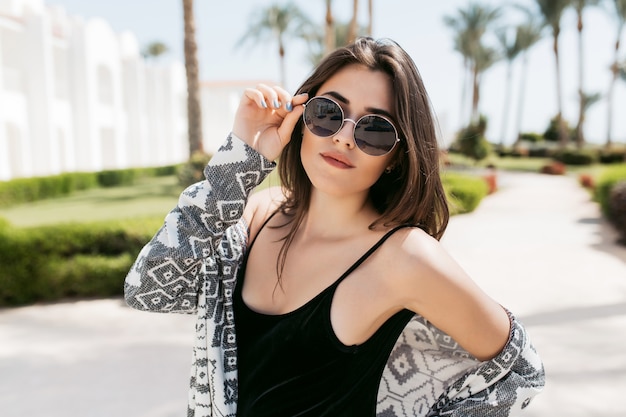 Portrait de gros plan de fille à la mode portant des lunettes de soleil et une chemise élégante, posant dans le parc et profitant du soleil du sud en vacances. Incroyable jeune femme aux cheveux raides foncés debout à l'extérieur.