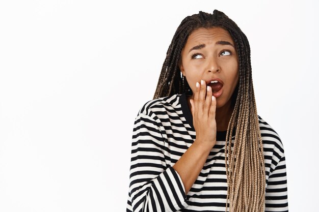 Portrait en gros plan d'une fille noire bâillant regardant de côté avec une expression de visage ennuyée non amusée debout dans un chemisier rayé sur fond blanc