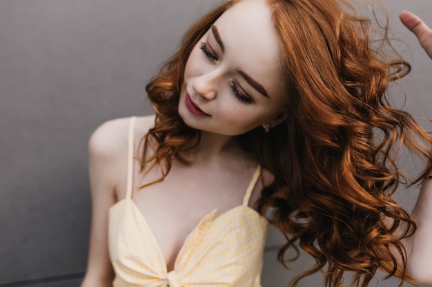 Portrait en gros plan d'une fille rousse sensuelle se détendre pendant la séance photo. Photo de merveilleuse dame frisée à la peau pâle isolée sur un mur gris.