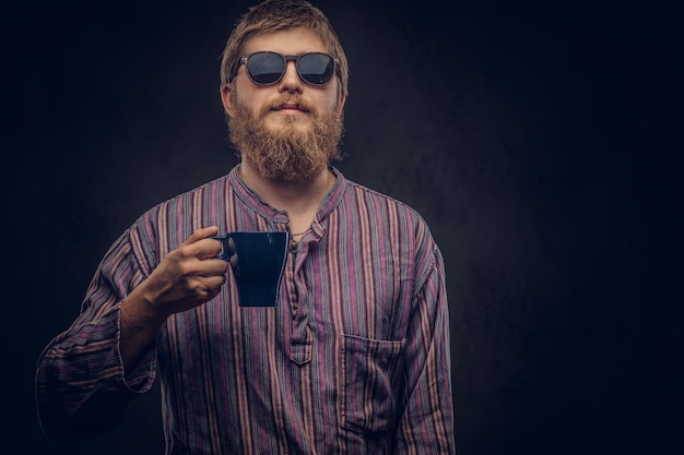 Portrait en gros plan d'un hipster portant des lunettes de soleil vêtu d'une chemise à l'ancienne tenant une tasse de café. Isolé sur un fond sombre.