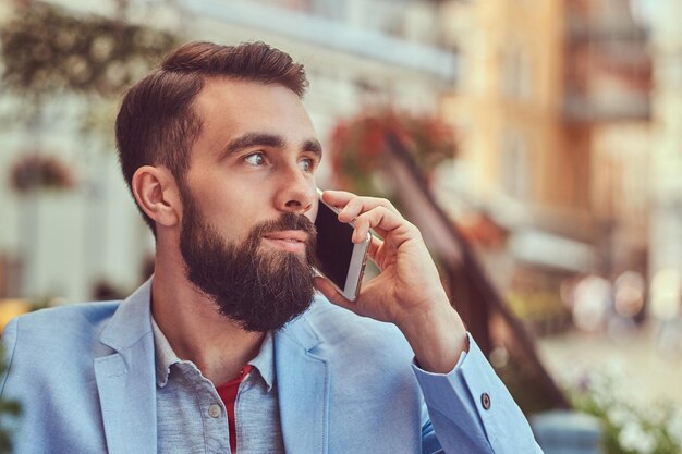 Portrait en gros plan d'un homme d'affaires barbu à la mode avec une coupe de cheveux élégante, parlant par téléphone, boit un verre de jus frais, assis dans un café à l'extérieur.