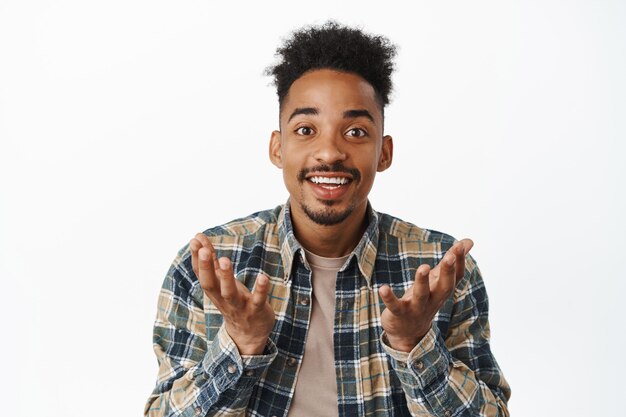 Portrait en gros plan d'un homme afro-américain heureux qui a l'air surpris et fier de vous, levant les mains comme s'il tenait qch dans les paumes vides, faisant une annonce, fond blanc