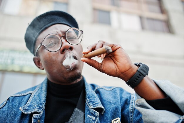 Portrait en gros plan d'un homme afro-américain en veste de jeans béret et lunettes fumant un cigare