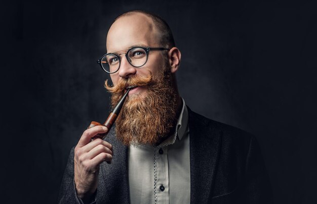 Portrait en gros plan d'un homme aristocratique à tête rasée dans des lunettes fumant une pipe sur fond gris.