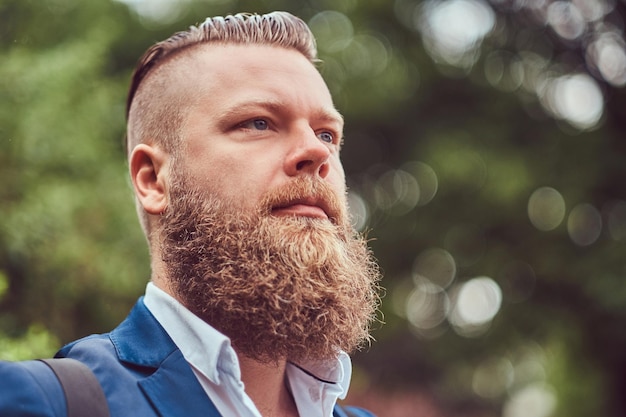 Portrait en gros plan d'un homme barbu avec une coupe de cheveux vêtu d'une chemise et d'une veste avec un sac à dos, debout dans un parc.