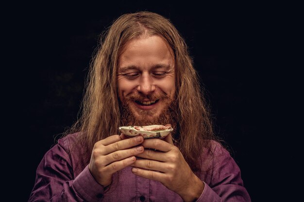 Portrait en gros plan d'un homme hipster rousse heureux aux cheveux longs et à la barbe pleine vêtu d'une chemise violette tenant une petite soucoupe avec du thé. Isolé sur fond texturé sombre.