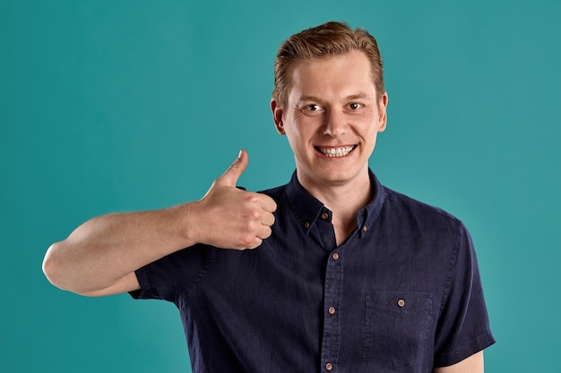 Photo gratuite portrait en gros plan d'un jeune beau gingembre dans un t-shirt bleu marine élégant regardant la caméra et montrant le pouce vers le haut tout en posant sur fond de studio bleu. expressions faciales humaines. émotions sincères