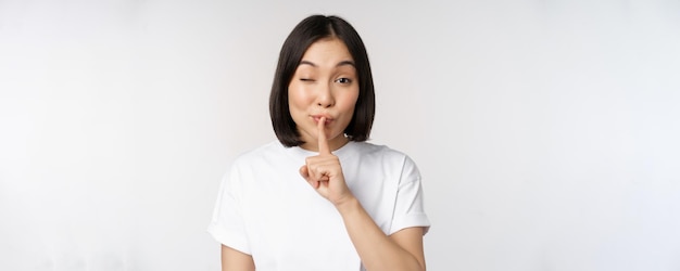 Portrait en gros plan d'une jeune belle fille asiatique qui chut a un geste secret garder le silence silencieux appuyez sur le doigt sur les lèvres debout en t-shirt sur fond blanc