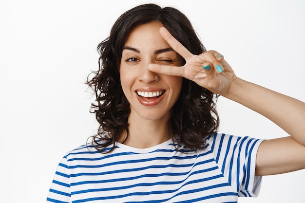 Portrait en gros plan d'une jeune femme heureuse, faisant un clin d'œil et montrant le geste du signe v de la paix, signe kawaii, dents blanches souriantes, debout gai sur blanc