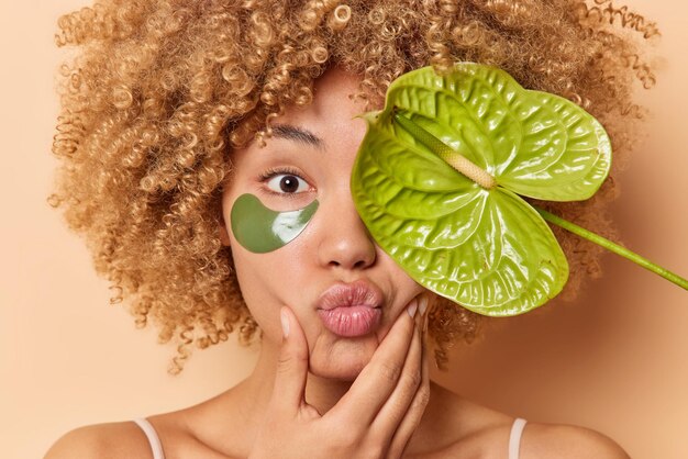 Portrait en gros plan d'une jeune femme à la peau soyeuse et saine garde les lèvres arrondies couvre les yeux avec la fleur de calla applique des patchs de collagène pour réduire les rides a des cheveux bouclés et touffus pose à l'intérieur. Le bien-être