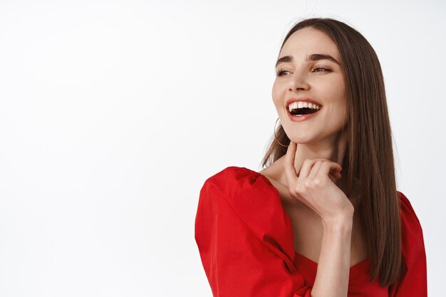 Portrait en gros plan d'une jeune femme séduisante en robe rouge, maquillage de soirée, rire et sourire heureux, profiter de la fête, regarder de côté la bannière du logo, lire le texte promotionnel, fond blanc