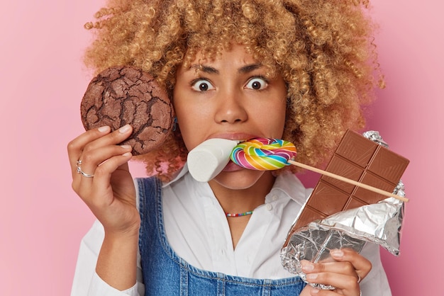 Portrait en gros plan d'une jeune femme sérieuse aux cheveux bouclés mange divers bonbons a la bouche pleine d'aliments nocifs concentrés dans la caméra porte une chemise blanche isolée sur fond rose Concept de vie douce