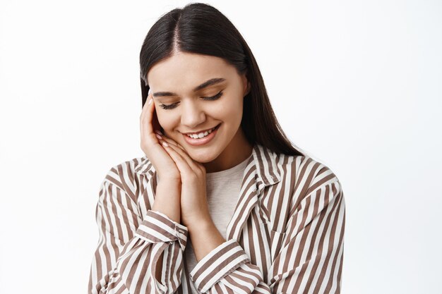 Portrait en gros plan d'une jeune femme tendre et féminine, regardant vers le bas et touchant une peau douce et propre avec un maquillage à la lumière naturelle, debout sur un mur blanc