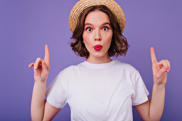 Portrait de gros plan d'une jeune fille aux yeux noirs surpris en chapeau d'été. Plan intérieur d'un modèle féminin bouclé drôle en t-shirt blanc posant avec les doigts sur le mur violet.
