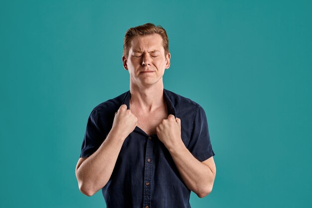 Portrait en gros plan d'un jeune homme athlétique au gingembre dans un t-shirt bleu marine élégant qui a l'air déçu tout en posant sur fond de studio bleu. Expressions faciales humaines. Concept d'émotions sincères. Copiez l'espace.