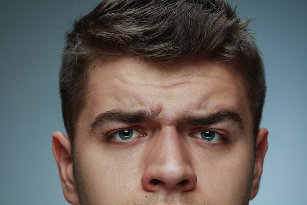 Portrait de gros plan de jeune homme isolé sur fond gris studio. Visage et yeux bleus du modèle masculin de race blanche. Concept de la santé et de la beauté des hommes, des soins personnels, des soins du corps et de la peau. En colère, a des rides.