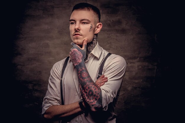 Portrait en gros plan d'un jeune tatoué à l'ancienne réfléchie portant une chemise blanche et des bretelles tient la main sur le menton. Isolé sur un fond sombre.