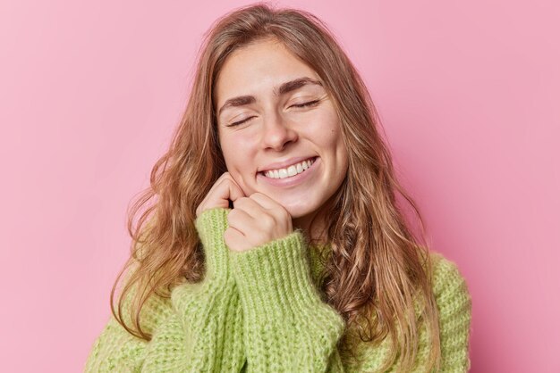 Portrait en gros plan d'une jolie femme heureuse sourit les yeux fermés garde les mains près du visage rêvasse à propos de quelque chose porte un pull vert tricoté isolé sur fond rose. Sentiments sincères