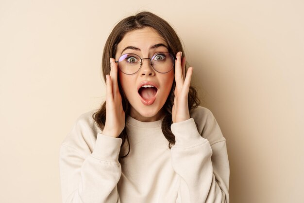 Portrait en gros plan d'une jolie femme à lunettes qui a l'air impressionnée, réagissant étonnée à ce qu'elle voit, debout sur fond beige.