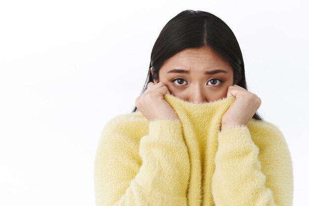 Portrait en gros plan d'une jolie fille asiatique timide et sombre fronçant les sourcils en tirant sur le visage et regardant la caméra effrayée tremblant de peur d'être en insécurité ou anxieuse debout effrayée sur fond blanc