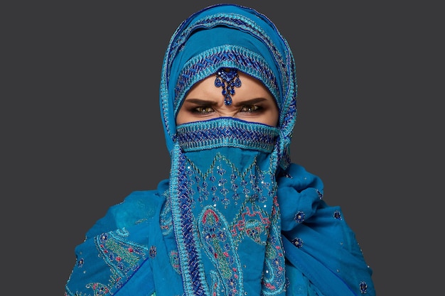 Portrait en gros plan d'une jolie fille aux charmants yeux charbonneux portant un élégant hijab bleu orné de paillettes et de bijoux. Elle pose et fait des grimaces sur un fond sombre. Émotions humaines, faciales