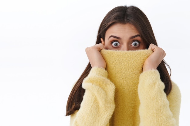 Portrait en gros plan d'une jolie fille surprise et surprise, se cachant le visage dans le col du pull en le tirant sur le nez et en exprimant le choc avec les yeux regardant la caméra sans voix effrayée ou effrayée sur fond blanc