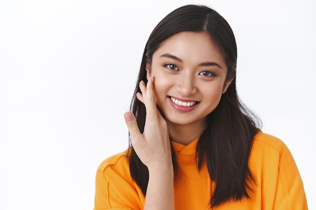 Portrait en gros plan jolie jeune femme asiatique touchant la joue et souriante, n'a pas de défauts, applique des produits de soin de la peau, maquillage coréen, mur blanc debout