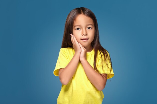 Portrait en gros plan d'une jolie petite fille joyeuse et mignonne avec une excellente peau et un sourire rayonnant, elle admire sa beauté dans un miroir, sur fond bleu