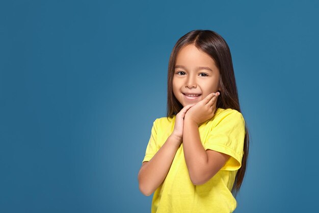 Portrait en gros plan d'une jolie petite fille joyeuse et mignonne avec une excellente peau et un sourire rayonnant, elle admire sa beauté dans un miroir, sur fond bleu