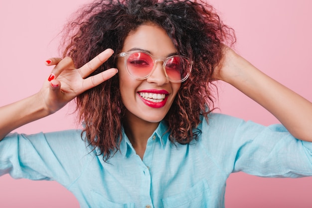 Portrait de gros plan de joyeuse fille africaine exprimant vraiment le bonheur. jolie dame mulâtre dans des verres roses montrant le signe de la paix.