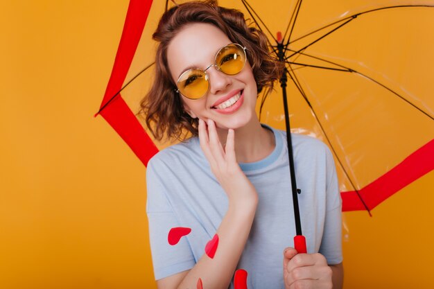 Portrait en gros plan d'une joyeuse fille aux cheveux bruns en t-shirt posant sous un parasol. Rire belle dame à lunettes de soleil bénéficiant d'une séance photo avec un parapluie.