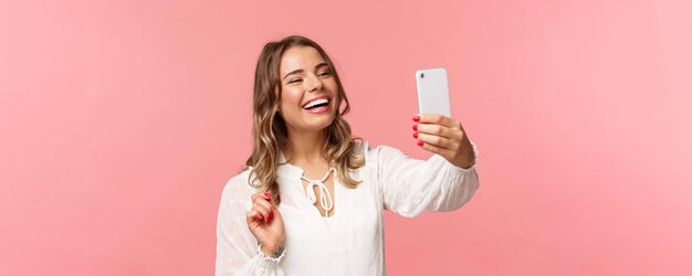 Portrait en gros plan d'une joyeuse fille blonde souriante et optimiste vêtue d'une robe blanche riant comme enregistrement vidéo appelant un ami sur une application mobile prenant un selfie photo sur fond rose de smartphone