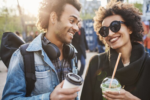 Portrait de gros plan de joyeux jeune couple d'amoureux tenant des boissons et souriant les uns aux autres tout en marchant dans le parc et en étant de bonne humeur