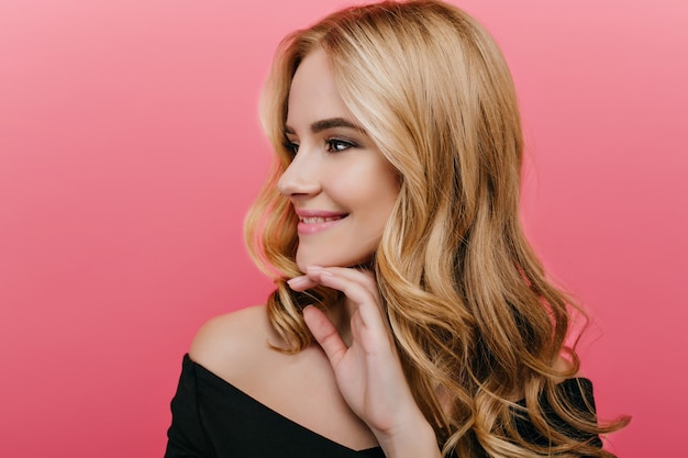 Portrait de gros plan de la magnifique jeune femme aux cheveux brillants isolé sur un mur rose. Photo intérieure d'une fille extatique aux cheveux blonds regardant ailleurs avec un sourire doux.