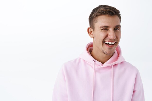 Portrait en gros plan d'un mec blond effronté en sweat à capuche rose, clin d'œil et rire, souriant largement, laissant entendre que vous aimez ce produit