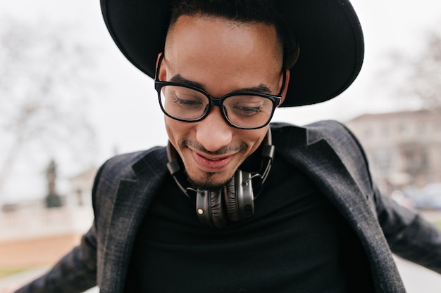 Portrait En Gros Plan D'un Mec Excité à La Peau Foncée Dansant Dans La Rue. Photo Extérieure D'un Modèle Masculin Bien Habillé En Chapeau Et Casque