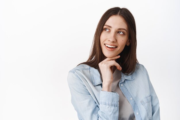 Portrait en gros plan d'un modèle féminin élégant et réfléchi, toucher le menton et regarder le coin supérieur gauche pensif, voir l'offre promotionnelle intéressante, lire la bannière du logo, debout sur un mur blanc