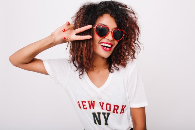 Portrait de gros plan de modèle féminin noir attrayant avec manucure rouge isolé. Photo d'une fille africaine heureuse à lunettes de soleil posant avec signe de paix.